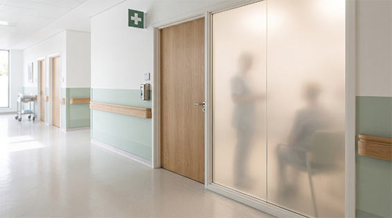 A hospital corridor beside an examination room with frosted glass walls, the blurred silhouettes of people inside just visible through the glass